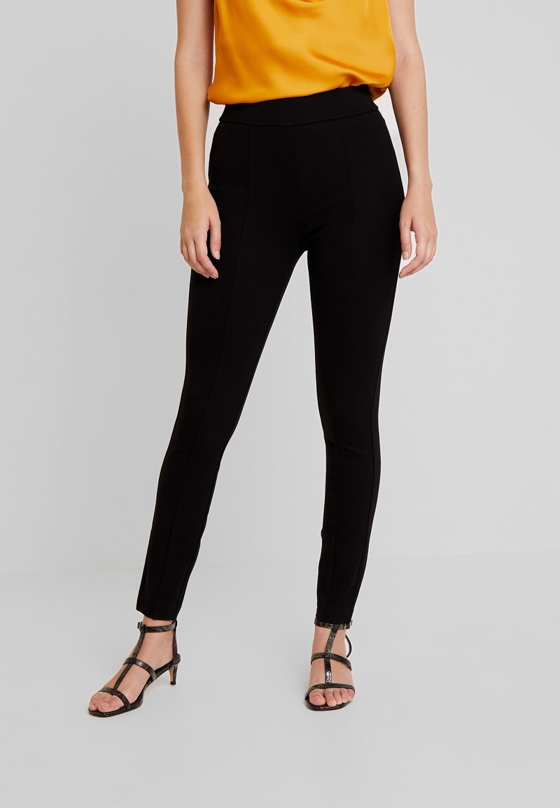 Černé Legíny s vysokým pasem, s přiléhavým střihem a vertikálními švy, doplněné černými sandály na podpatku a žlutým topem.