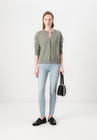 Light green, fuzzy cardigan with a round neckline and button closure, paired with light blue skinny jeans and black loafers, holding a black bag.