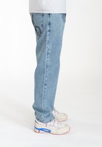 Jean en denim bleu clair avec des jambes droites, présentant un design distinctif des poches arrière. Portés avec des chaussures de sport blanches avec des accents colorés.