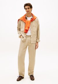 Beige Baumwolljacke mit Knopfverschluss, kombiniert mit passenden beigen Hosen. Ein orangefarbenes Sweatshirt hängt um den Hals. Braune Schuhe.