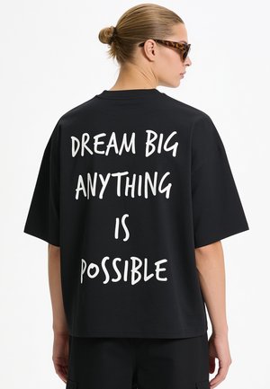 Frau mit Dutt und Schildpattsonnenbrille trägt schwarzes Shirt mit weißem Text "Dream big anything is possible" auf dem Rücken.