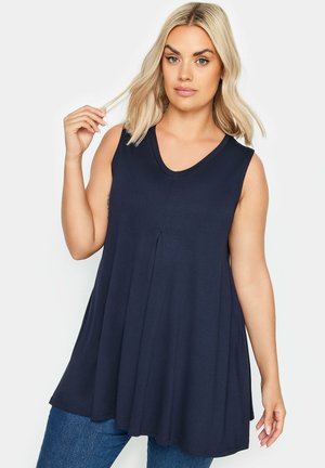 Lyshåret kvinde iført ærmeløs marineblå V-hals løs top og blå jeans, holder en hårtot med et neutralt udtryk.