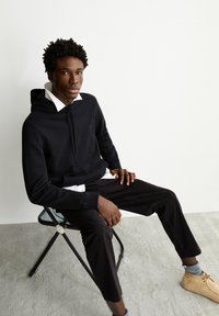 Zwarte hoodie met trekkoord, witte overhemd met kraag eronder, zwarte broek, zittend op een zwarte metalen stoel met een lichtgrijs tapijt. Licht schoeisel.