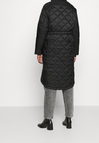 Abrigo largo negro acolchado con cintura ceñida, que presenta una textura suave y un patrón de diamantes. Combinado con jeans grises y botas de tobillo negras y brillantes.