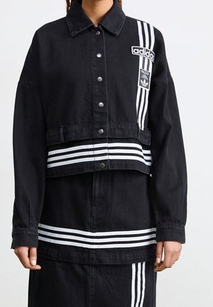 Persona vestida con un conjunto de chaqueta y falda negra de Adidas con rayas y logotipos blancos, con botones a presión y cuello, de pie contra un fondo liso.