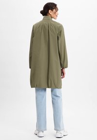 Olijfgroene, lange mouwen, knie-lange jas met hoge kraag, achterplooien en elastische zoom, gecombineerd met lichtblauwe jeans en witte schoenen.