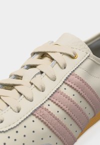 Κοντινό πλάνο μπεζ αθλητικού παπουτσιού με κρέμ κορδόνια και τρεις παράλληλες ροζ σκούρες σουέντ ρίγες σε διάτρητο δερμάτινο επάνω μέρος.