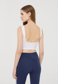 Top bianco corto con spalline larghe, schiena bassa, texture morbida, abbinato a leggings a vita alta blu scuro. Design semplice, senza motivi o accessori.