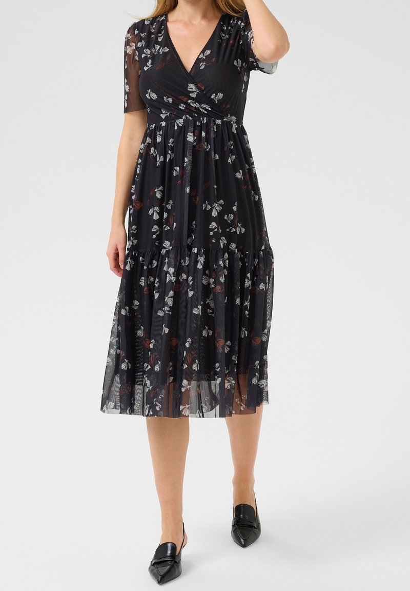 Femme portant une robe midi noire avec imprimé floral blanc et rouge et manches courtes transparentes, associée à des chaussures slingback noires à bouts pointus.