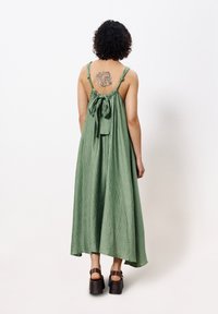 Langes, grünes Baumwollkleid mit strukturierter Oberfläche, verstellbaren Trägern und einem großen Schleifendetail auf dem Rücken. Kombiniert mit braunen, klobigen Sandalen.