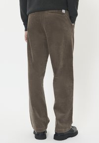 Bruine corduroy broek met een rechte snit, voorzien van een achterzak en een subtiele ribbelstructuur. Gepaarde met zwarte schoenen, op een effen achtergrond.