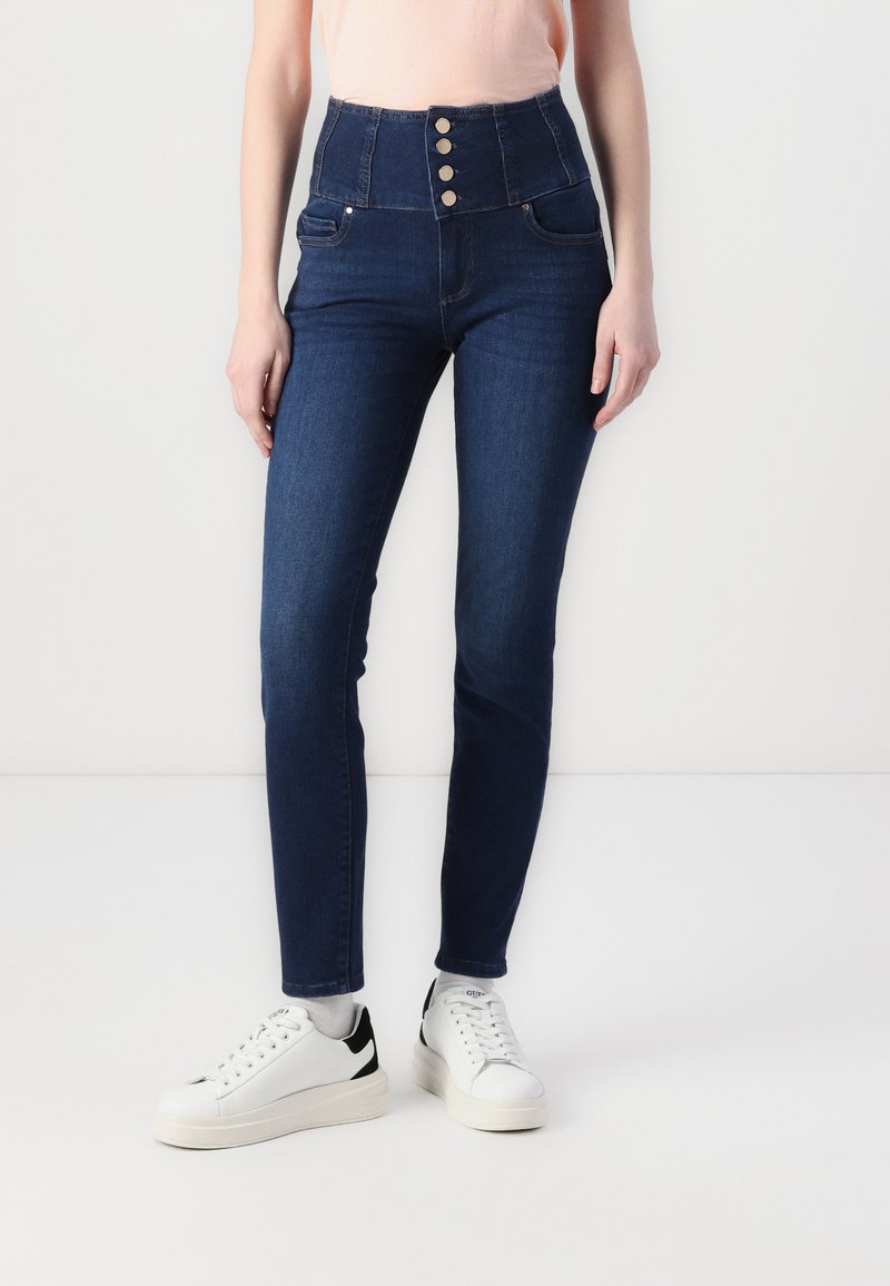 Jeans din denim de culoare albastră închisă, cu talie înaltă, cu închidere prin cinci butoane și buzunare frontale, asortați cu adidași albi, având un design elegant și ajustat.