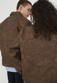 Jachete din piele ecologică maro, cu un mare design brodat pe spate, având o textură netedă și o croială relaxată.