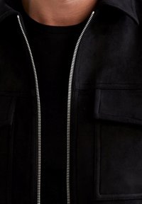 Giacca in suede nera con zip argentata, due tasche frontali, colletto e texture morbida. Indossata sopra una camicia nera.