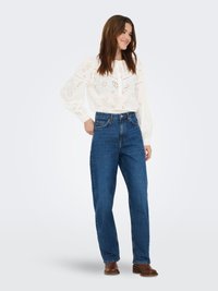 Blouse blanche en tissu ajouré avec des découpes florales, manches longues et col boutonné, associée à un jean en denim bleu taille haute et des bottines marron.