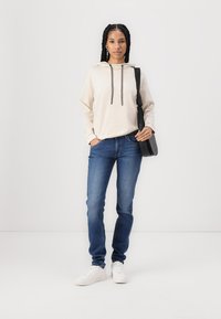 Camisola bege com cordões cinzentos, jeans de ganga azul e sapatilhas brancas. Apresenta uma mala de ombro preta e um penteado trançado.