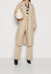 Abrigo de lana beige con cuello extragrande; superpuesto sobre un top corto estampado y pantalones beige de pierna ancha. Modelo sosteniendo un bolso crema. Botas negras con tacón.