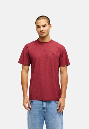 Jonge man staat met het gezicht naar voren, draagt een donkerrode korte mouwen T-shirt en lichtblauwe spijkerbroek tegen een effen lichte achtergrond.
