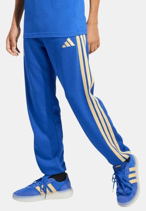 Person trägt blaue Adidas-Trainingshose und passende blaue Adidas-Sneaker mit beigen Streifen vor einfarbigem Hintergrund.