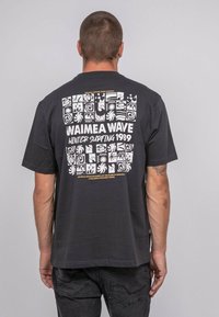 T-shirt noir à manches courtes présentant un design graphique blanc au dos avec des motifs abstraits et le texte "WAIMEA WAVE WINTER SURFING 1989."
