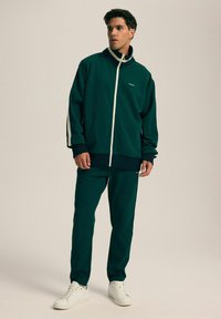 Ciemnozielony dres z białymi akcentami, z kurtką zapinaną na zamek i dopasowanymi spodniami, wykonany z gładkiego materiału o luźnym kroju.