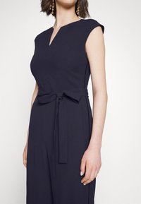 Žena v černém overal s bezrukávovým střihem, s výstřihem do V a látkovým opaskem uvázaným do mašle v pase, s zlatými náušnicemi.