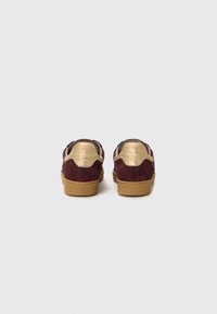 Zapatillas de ante burdeos con acentos dorados, panel trasero texturizado y suela de goma color marrón claro, con forma redondeada y detalles de costura.