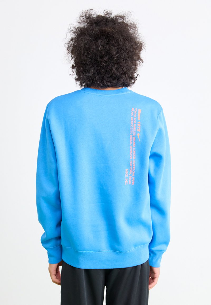 Blå sweatshirt lavet af bomuldsblanding, med rund hals og lange ærmer. Tekst i orange på ryggen indeholder forskellige bynavne.