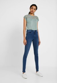 Top curto de teal claro com detalhes em renda nos ombros, combinado com jeans skinny azul escuro e ténis brancos. Design simples, look casual.