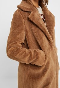 Mujer con un abrigo de teddy marrón con solapas anchas y manos en los bolsillos, visible un suéter blanco y con patrones debajo.