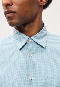 Ανοιχτό μπλε καρό πουκάμισο με απαλή υφή, κουμπωτό γιακά και αντίθεση στα κουμπιά. Διαθέτει κεντημένο λογότυπο στην τσέπη του στήθους.