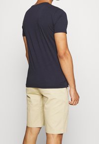 Námořnická modrá bavlněná tričko s krátkým rukávem, kulatým výstřihem, nošené s světle béžovými šortkami. Hladká textura s klasickým střihem a jednoduchým designem.