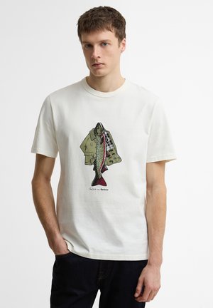 Νεαρός άνδρας που φοράει λευκό T-shirt με γραφικό ενός ψαριού που συγχωνεύεται σε ένα πράσινο σακάκι μπροστά, συνδυασμένο με σκούρο παντελόνι.