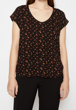 Kobieta w czarnej bluzce z krótkim rękawem i dekoltem w serek, z drobnym czerwonym, różowym i żółtym kwiatowym wzorem, do tego czarne spodnie.