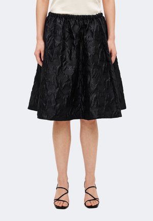 Jupe noire longueur genou avec motif floral texturé portée avec un top blanc et des sandales à talons hauts noires à brides sur un fond clair.