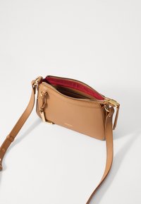 Borsa a tracolla in pelle tan con finitura testurizzata, hardware color oro e interno rosso. Presenta una chiusura con zip e una tracolla regolabile.