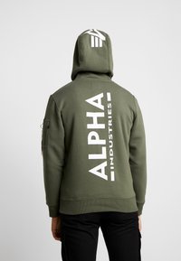 Olivengrønn hettegenser med et stort hvitt "ALPHA INDUSTRIES"-logo på ryggen, justerbar hette og glidelås-lomme på armen. Blandingsmateriale av bomull.