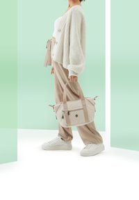 Beige håndtaske med mønstret stof, dobbelte øverste håndtag og lynlåse, holdt af en person iført en cremefarvet cardigan og lyse bukser.