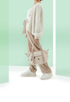 Beigen värinen käsilaukku, jossa on kuvioitu kangaspinta, kaksi yläkahvaa ja vetoketjut, henkilö pitää sitä kädessään yllään kermaisen värinen neuletakki ja vaaleat housut.