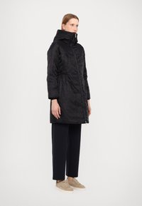 Cappotto trapuntato nero con colletto alto, zip frontale, vita elastica e maniche lunghe, abbinato a pantaloni scuri e sneakers chiare.