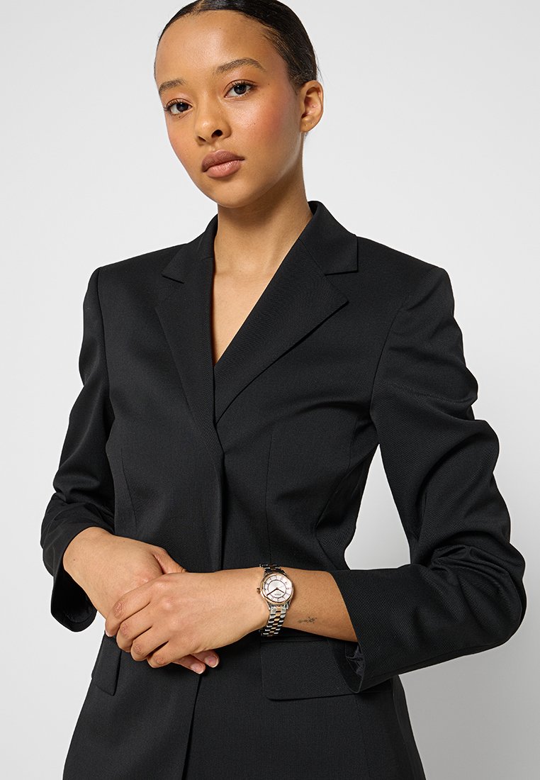 Veste de costume noire avec des épaules structurées, un tissu texturé, une coupe ajustée et des revers en pointe. Le modèle porte une montre en argent et or rose.