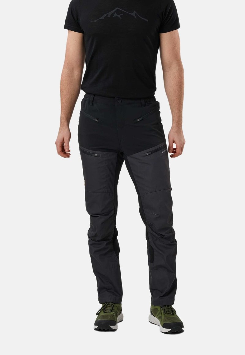 Homme portant un pantalon d'extérieur noir avec des poches zippées et des chaussures de randonnée vertes, debout devant un fond uni.