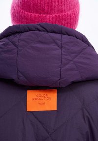 Nafouknutá fialová bunda s prošívaným vzorem, s oranžovou nálepkou na zadní straně, na které je červeným písmem nápis "COLOR REVOLUTION".