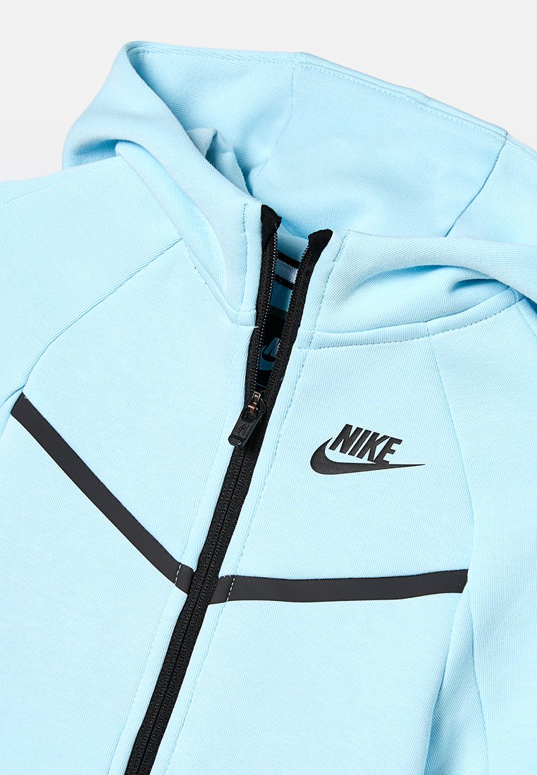 Haut de sweatshirt zippé bleu clair avec une fermeture éclair noire et un logo noir. Le tissu a une texture lisse avec des rayures noires contrastantes sur la poitrine.