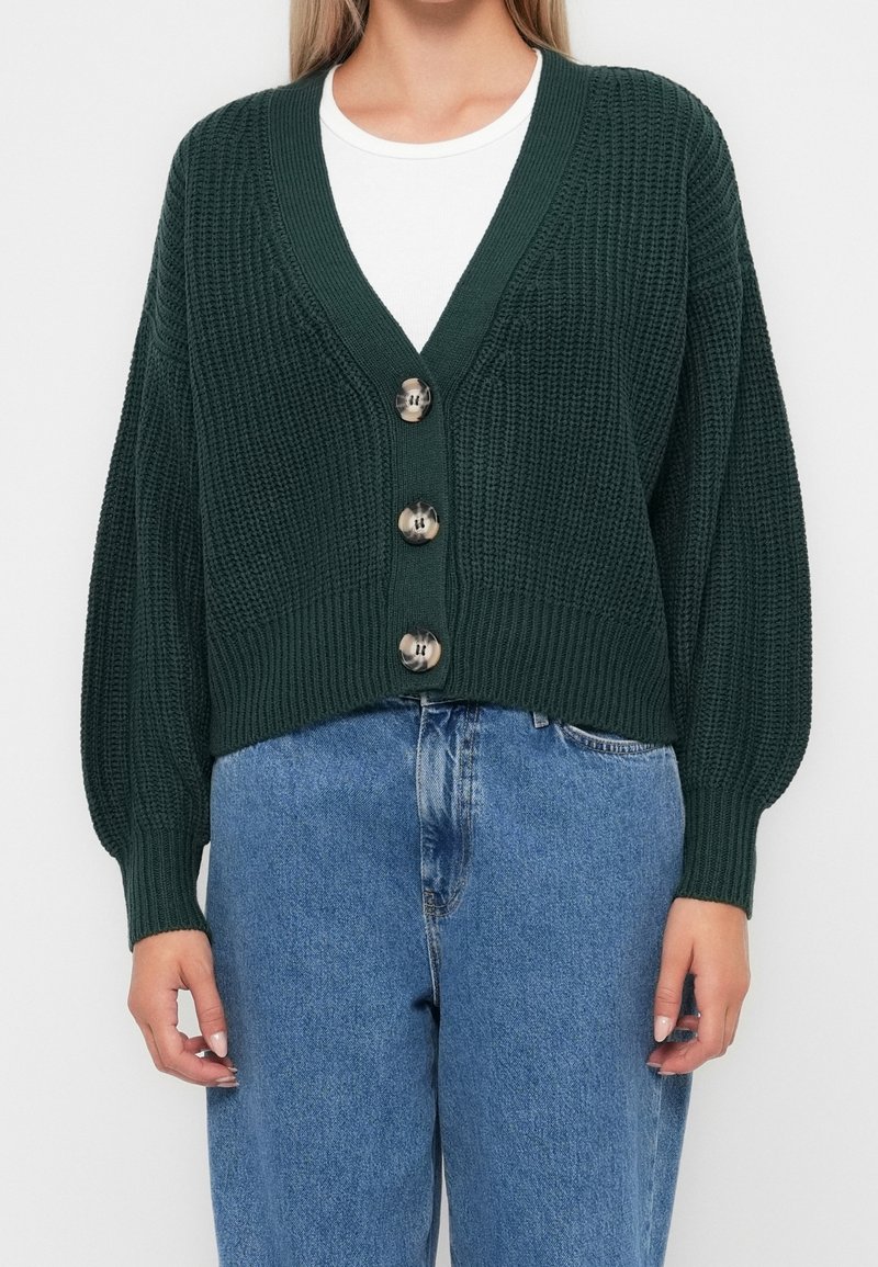 Femme portant un cardigan en maille épaisse vert foncé avec trois gros boutons sur un top blanc, associé à un jean bleu.