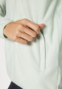 Hele roheline fliistekott, millel on eesmine tasku, kus on kuvatud lähedalt käsi, mis haarab tasku avamisest, rõhutades pehmet tekstuuri ja materjali.