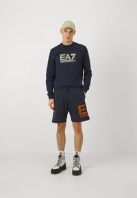 Felpa navy con logo EA7 bianco e argento, abbinata a pantaloni corti navy con logo EA arancione. Indossata con stivaletti bianchi con lacci.