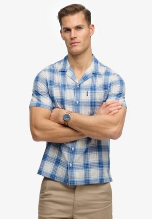 Chemise à manches courtes à carreaux bleus et blancs, avec un devant boutonné, un tissu léger et un col. Portée avec un pantalon beige.