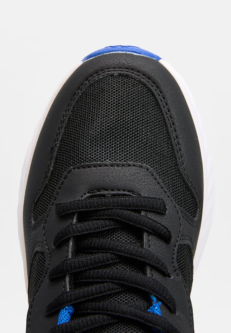 Zapatilla negra con malla y materiales sintéticos, con acentos azules, cordones texturizados y una suela blanca, destacando un diseño deportivo.