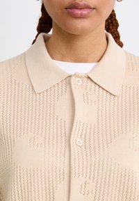 Beige knitted cardigan with a collared neckline, featuring openwork texture, two buttons, and subtle patterning across the fabric.